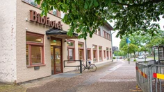 Kinna bibliotek, en byggnad i ljus tegel med texten Bibliotek ovanför glasdörren i entrén. En cykel står parkerad på gatan utanför och ett stort träd breder ut sina grenar framför huset.