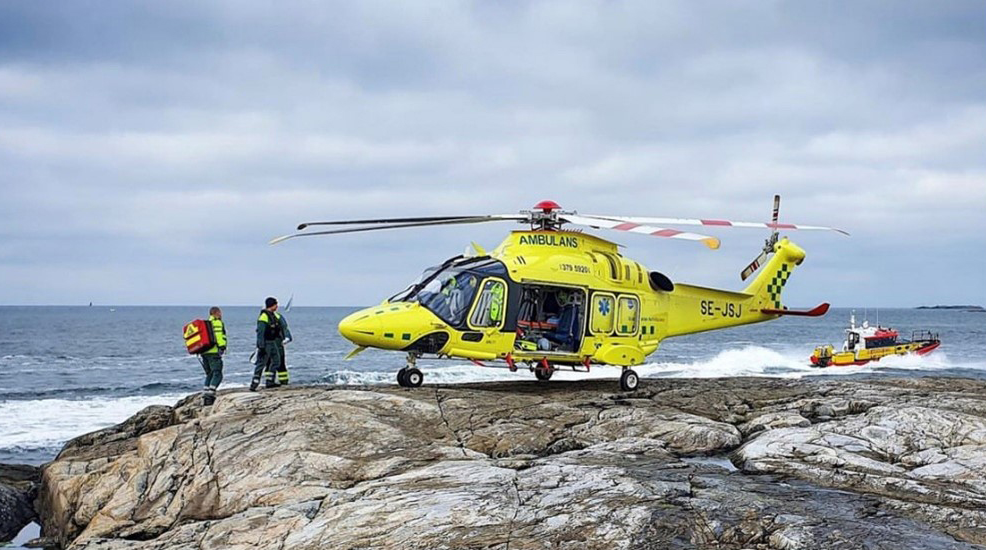 En ambulanshelikopter står parkerad på klippor vid havet. Sjukvårdspersonal står runt den och i bakgrunden syns en båt.