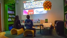 Två barn sitter på varsin pall i ett mörkt rum. På väggen framför dem är ett tv-spel projicerat. De håller i varsin spelkontroll och har blicken fäst på spelet på väggen. Två gröna bokhyllor står utmed väggarna och ett tråg med barnböcker står under spelet på väggen. På golvet ligger en grön mjuk matta.