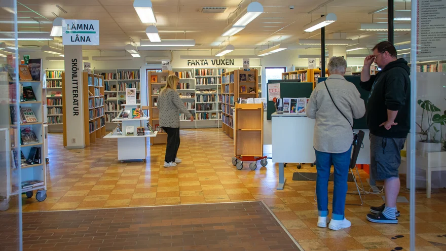 I entrén till Sotenäs kommunbibliotek i Kungshamn. Biblioteksassistent Emil Andersson hjälper en besökare med något, en kvinnlig bibliotekarie är på väg i rummet. Golvet har en gulrutig plastmatta och på skyltar i rummet står bland annat "Lämna", "Låna" och "Skönlitteratur".