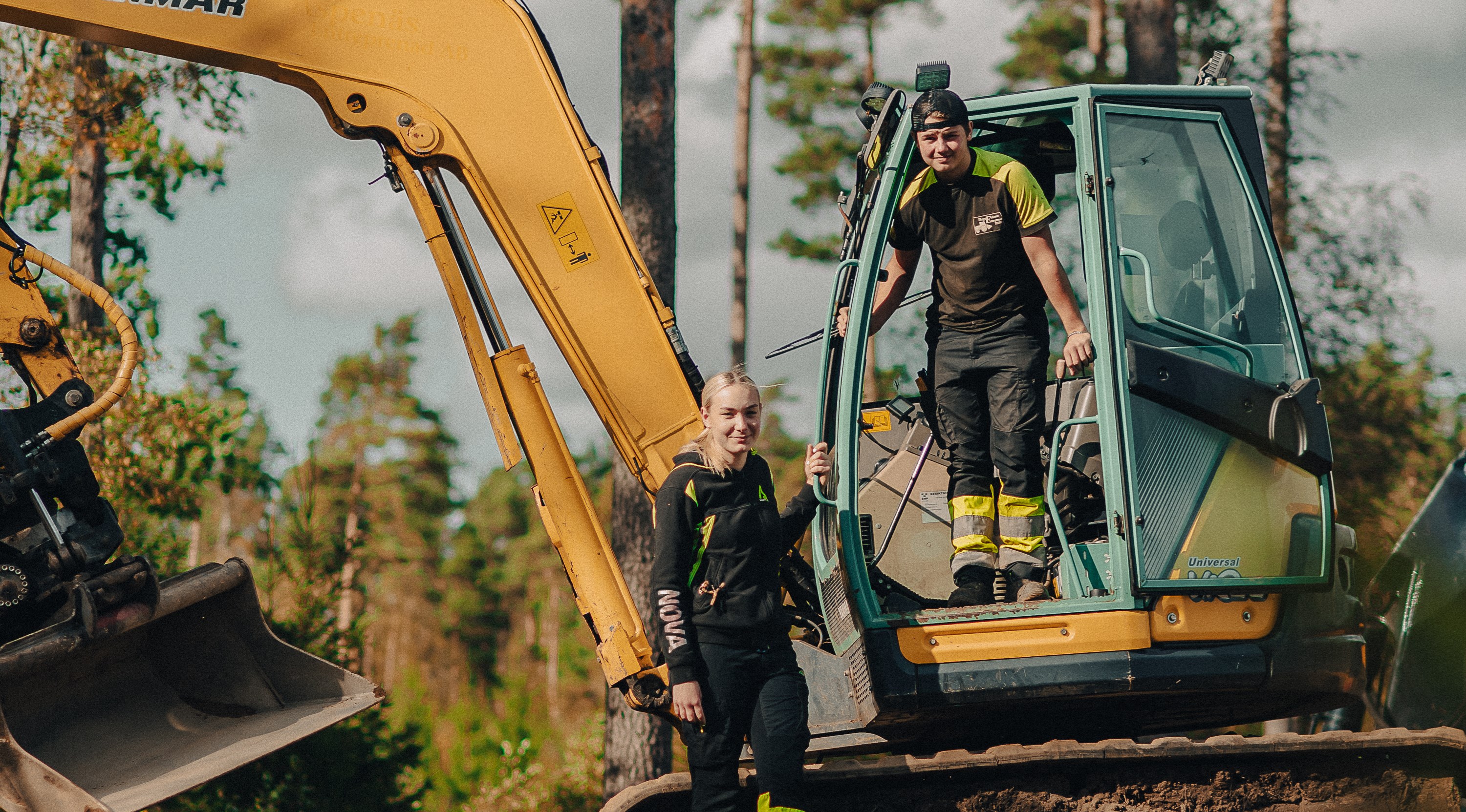 Två elever står vid grävmaskin i skogen