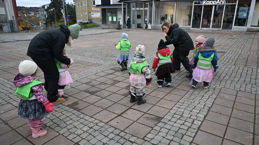 Sju förskolebarn går omkring på ett torg med två pedagoger.
