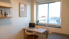 Möblerat internatrum med stort fönster, skrivbord, säng och vägghylla