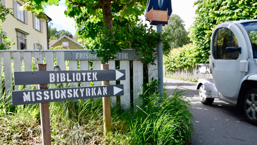 En golfbil susar förbi på en smal väg. Två träskyltar visar vägen, på den ena står det Bibliotek, på den andra Missionskyrkan.