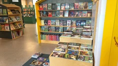 Bokexponering i biblioteksrummet. Böcker står uppställda med framsidan utåt på hyllor och ligger på bord i olika höjd. Bokhyllorna är utformade som små huskroppar och slottstorn. Färgerna i rummet går i gult och dovt grön.