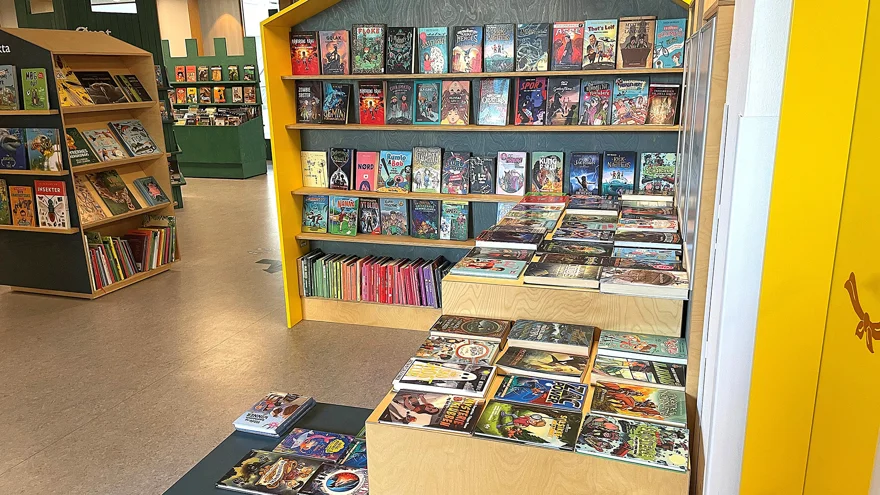 Bokexponering i biblioteksrummet. Böcker står uppställda med framsidan utåt på hyllor och ligger på bord i olika höjd. Bokhyllorna är utformade som små huskroppar och slottstorn. Färgerna i rummet går i gult och dovt grön.